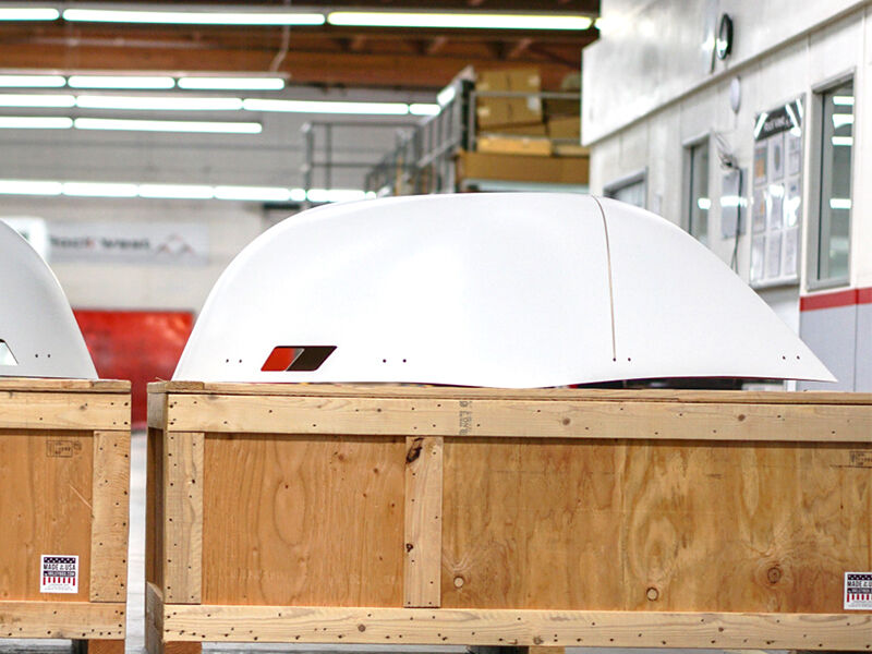 Image of the X-band radome sitting on a wooden shipping crate in a manufacturing environment. The radome is painted white and has sets of machine holes and a cut out near the base.